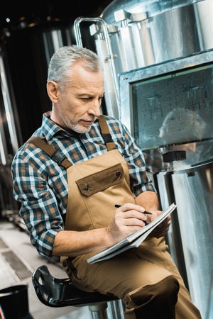 senior brewer in working overalls writing in notepad while examining brewery equipmentの写真素材