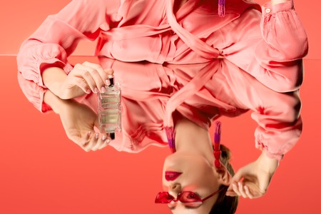cropped view of woman in blouse and red sunglasses posing with perfume bottle and mirror reflection...