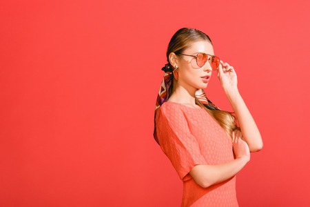 fashionable girl posing in living coral dress and sunglasses isolated on red background