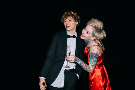 cheerful woman in red dress standing with boyfriend in suit and holding glass with drink isolated on blackの写真素材