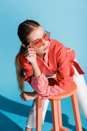 stylish woman in living coral clothing and sunglasses posing on stool on blueの写真素材