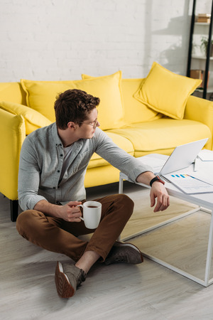 tired man in glasses sitting on floor and holding cup with drink at homeの写真素材