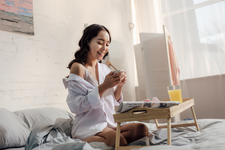 happy seductive asian girl in white shirt holding cup of coffee and having breakfast on bedの写真素材