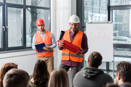 selective focus of handsome firemen in helmets standing near white board with fire safety lettering and holding extinguisher and clipboard in handsの写真素材