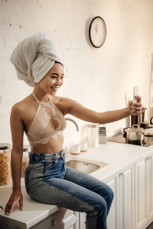 attractive smiling young woman with towel on head taking selfie with smartphone in kitchenの写真素材