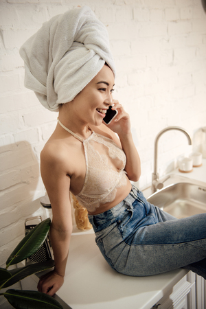 attractive asian woman with towel on head talking by smartphone while sitting in kitchenの写真素材