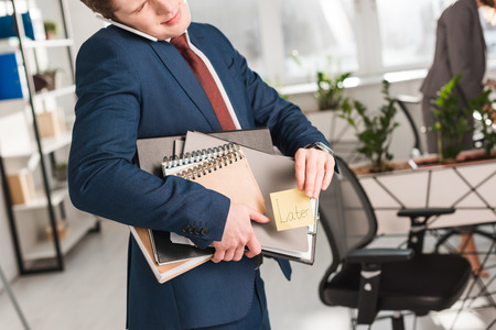 cropped view of businessman holding folders and sticky note with later lettering and talking on smartphone in officeの写真素材