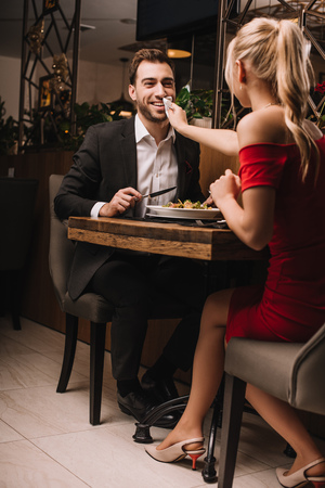 blonde girlfriend wiping mouth of smiling boyfriend with napkin in restaurantの写真素材