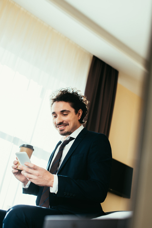 smiling businessman using smartphone and holding coffee to go in hotel roomの写真素材