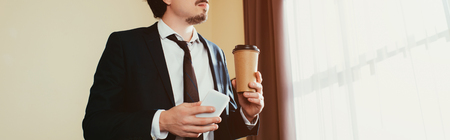 cropped view of businessman using smartphone and holding coffee to go in hotel room with sunlightの写真素材