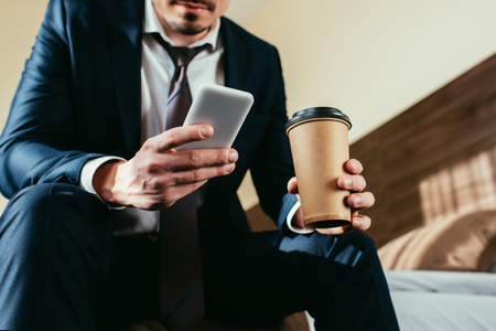 cropped view of businessman using smartphone and holding coffee to go in hotel roomの写真素材