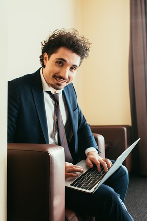 smiling businessman in suit working on laptop in hotel room