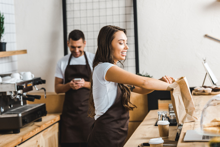 attractive cashier in brown apron packing paper bag wile barista looking to smartphone near bar counter in coffee houseの写真素材