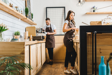 attractive cashier with paper bag and barista making coffee behind bar counter in coffee houseの写真素材
