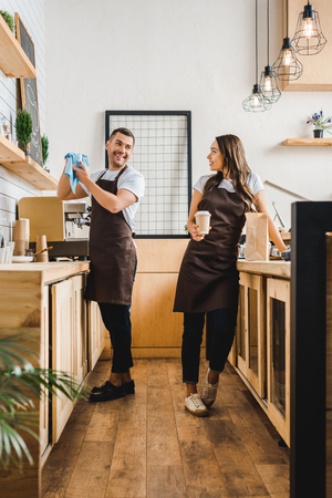 barista and cashier in aprons standing and talking behind bar counter in coffee houseの写真素材