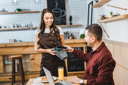 attractive waitress holding terminal wile handsome freelancer sitting at table with laptop and paying with smartphone in coffee houseの写真素材