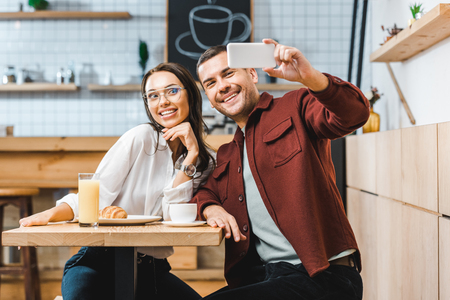 attractive brunette woman and handsome man in burgundy shirt sitting at table and taking selfie to smartphone in coffee houseの写真素材