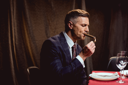 handsome man in suit sitting at table and lighting up cigar in restaurantの写真素材