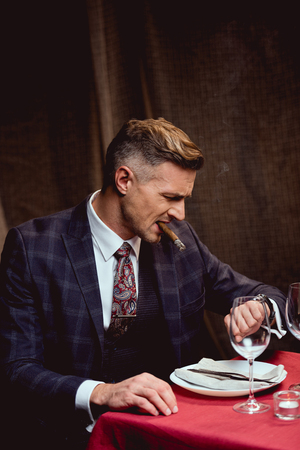 dissatisfied handsome man in suit sitting at table, smoking cigar and looking at watch in restaurantの写真素材