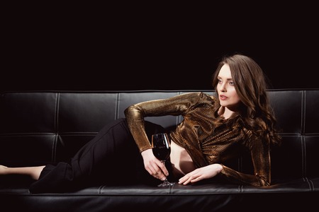 beautiful elegant woman lying on couch with glass of red wine isolated on blackの写真素材