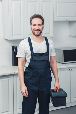 adult smiling bearded repairman in working overall holding toolbox in kitchen at homeの写真素材
