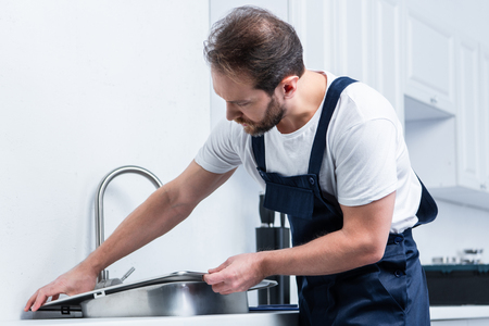adult bearded repairman in working overall fixing sink in kitchenの写真素材