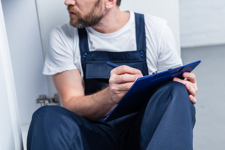 partial view of adult craftsman writing in clipboard while sitting on floor in kitchen