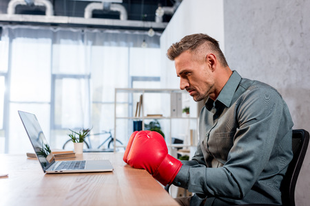 handsome businessman watching championship on laptop while wearing boxing gloves in officeの写真素材