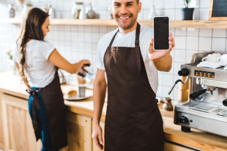 selective focus of handsome cashier showing smartphone with blank screen wile barista working in coffee houseの写真素材