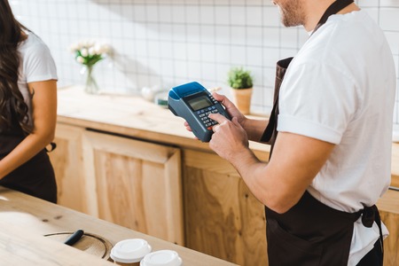 cropped view of cashier holding blue terminal wile brunette barista working in coffee houseの写真素材