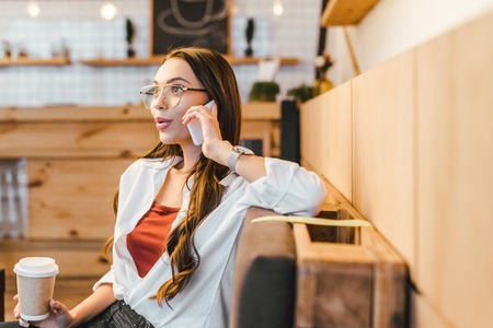 attractive woman in white shirt sitting on couch, holding paper cup and talking on smartphone in coffee houseの写真素材