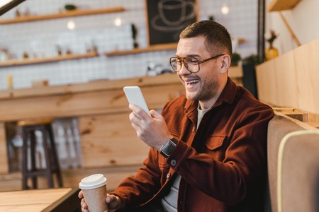 handsome man in burgundy shirt sitting on couch, holding paper cup, looking to smartphone and laughing in coffee houseの写真素材