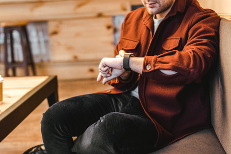 cropped view of man in burgundy shirt sitting on couch and touching watch in coffee house