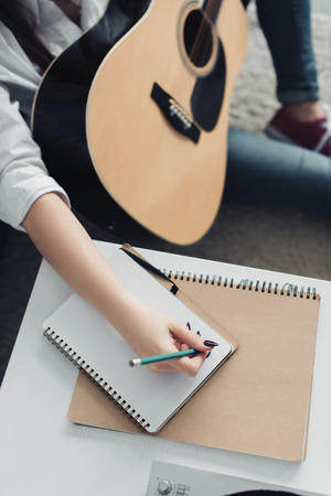cropped view of girl with acoustic guitar writing in notebook while composing music at homeの写真素材