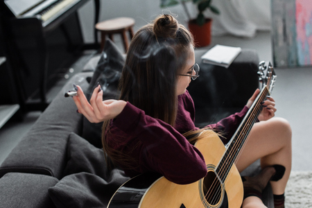 girl sitting, holding marijuana joint and playing guitar in living room