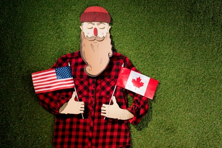 cardboard man in plaid shirt and hat holding canadian and american flags on green grass backgroundの写真素材