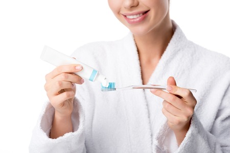 partial view of woman in white bathrobe putting toothpaste on toothbrush isolated on white
