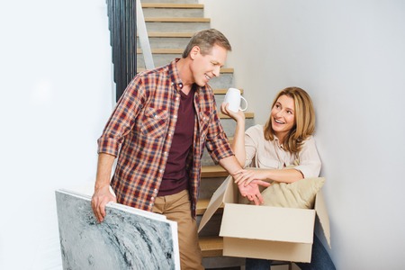 smiling man holding picture while standing near wife sitting on stairs and unpacking carton boxの写真素材
