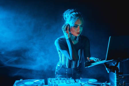 serious dj woman with blonde hair looking at laptop while listening music in headphones in nightclub...