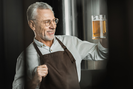 smiling male brewer looking at glass of beer in brewery