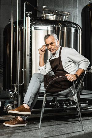 senior brewer in apron sitting on chair in breweryの写真素材