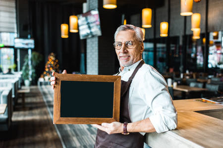 smiling owner of pub holding empty board and standing near bar counter