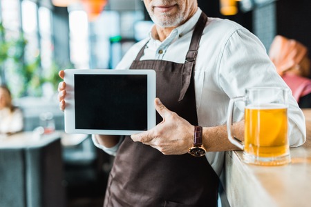 cropped view of male owner holding digital tablet with blank screen in pub with glass of beerの写真素材