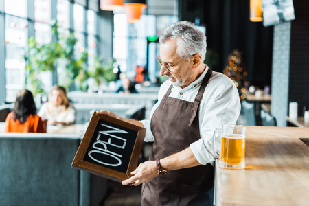 senior owner of pub holding open sign and standing near bar counter with glass of beerの写真素材