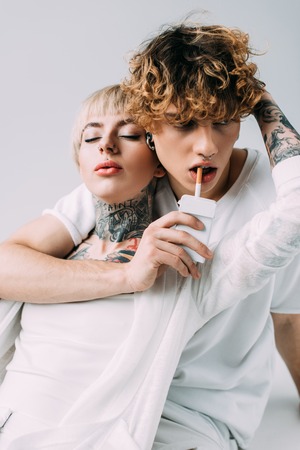 tattooed woman hugging handsome man with curly hair holding cigarette in mouth isolated on grey