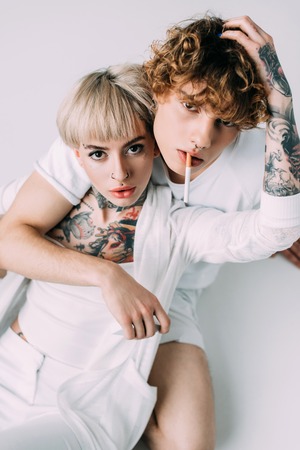 handsome man with curly hair holding cigarette in mouth while sitting with blonde tattooed girl...