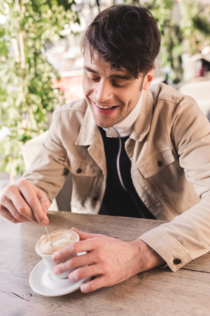 cheerful man holding spoon in cup with cappuccino in cafeの写真素材