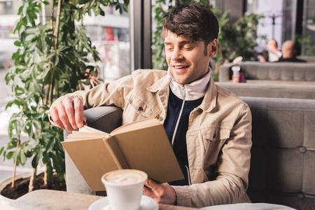 happy man holding book near cup with cappuccino in cafeの写真素材