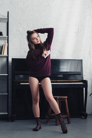 beautiful girl with headphones on head holding smartphone and standing near piano at homeの写真素材