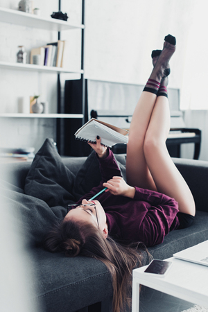 attractive girl lying on sofa and holding notebook and pencil in living roomの写真素材
